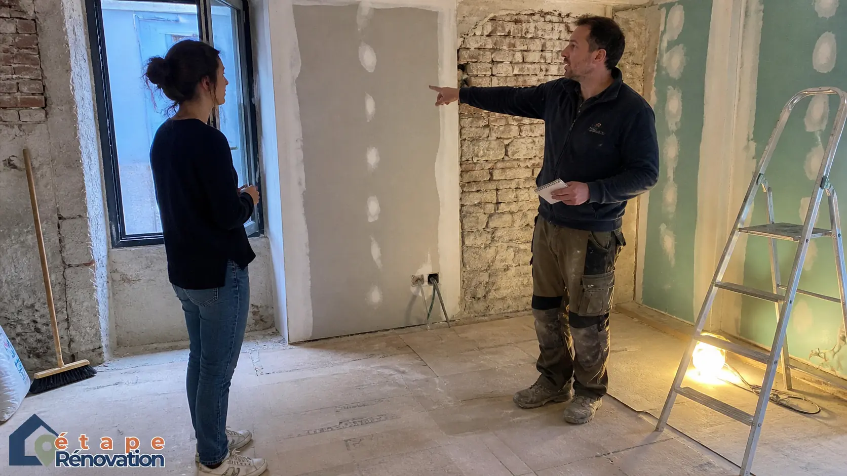 Photographie sur le vif d'une propriétaire et d'un maître d'œuvre discutant des travaux au milieu d'un salon en pleine rénovation avec murs à nu et plaques de plâtre.